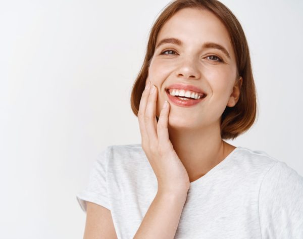 Beauty. Close-up of happy woman with short hair, touching clean fresh facial skin and smiling, showing perfect teeth and face, standing against white background.