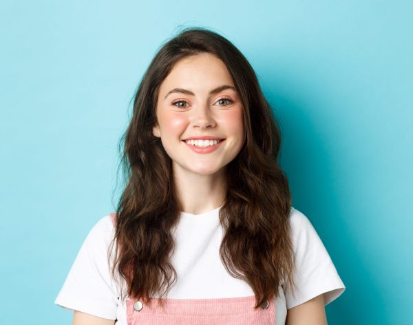 Close up portrait of cheerful glamour girl with cute make up, smiling white teeth and looking happy at camera, standing over blue background.